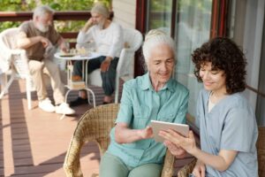 Healthcare worker showing elderly woman information on a tablet while two older adults play chess in the background on a sunny veranda
